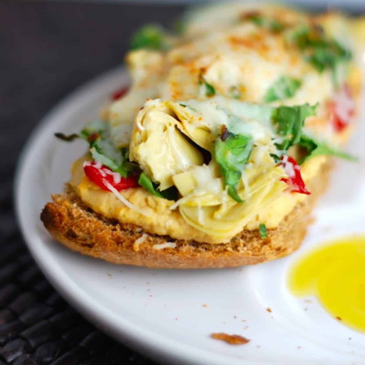 Ppen-faced sandwich with artichokes and red peppers over hummus and bread, drizzled with olive oil.
